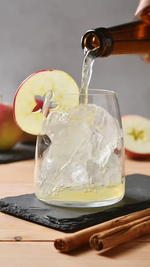 Apple cider pours over ice in glass closeup beside apple slices and cinnamon for Stock Footage 345558240