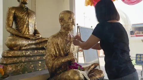 Applying gold leaf to Buddha statue making wishes for good fortune, representing Stock Footage 328492023