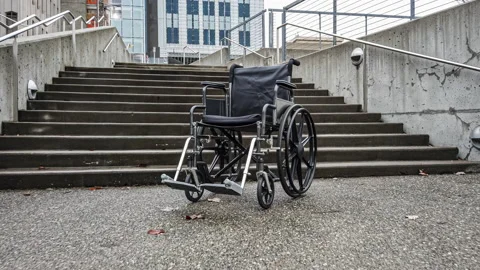 Approaching an empty wheelchair sitting at the base of concrete steps Stock Footage 218643182