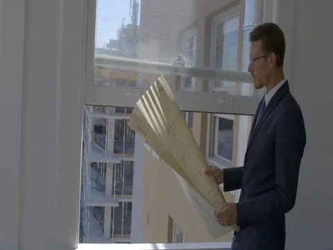 Architect with blueprint in front of window overlooking construction 4K Stock Footage 80232235