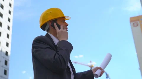 The architect in a yellow helmet stands at the construction site and talks on Vídeos de archivo 89175385