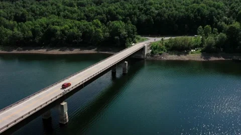 Ariel drone shot of red car driving across a bridge over a lake into the forest Stock Footage