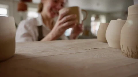 Artisan in Apron Joyfully Examining Handmade Mug in Ceramics Studio with Blurred Stock Footage 266693751