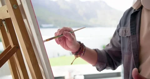 Artist is checking canvas and making short strokes for detail at easel by window Stock Footage 336247522