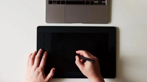 Artist's hands using a digital drawing tablet and stylus with a laptop nearby Stock Footage 337173698