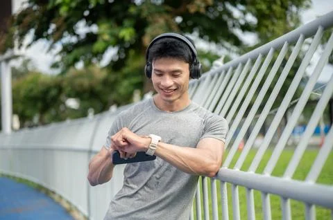 Asian athletic man smiling while checking time on smartwatch leaning on fen.. Photos
