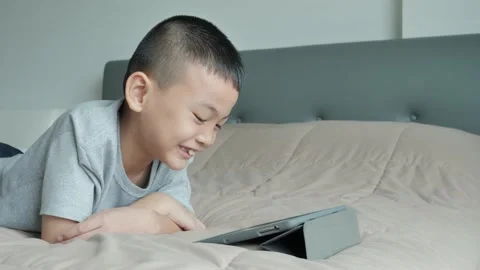 An Asian boy enjoys using a tablet computer on the bed in the bedroom. Stock Footage 141126497