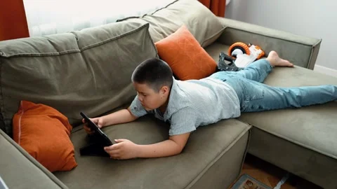Asian boy holds game controller on sofa, striped shirt and jeans, video game Video stock 312661569