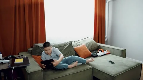 Asian boy holds game controller on sofa, striped shirt and jeans, video game 動画素材 312661655