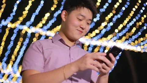 Asian boy typing in his smartphone and talking. Many emotions. Night time Stock Footage 93255042