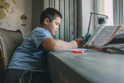 Asian boy write in notebook, homework beside open textbook on desk, study tool 库存照片