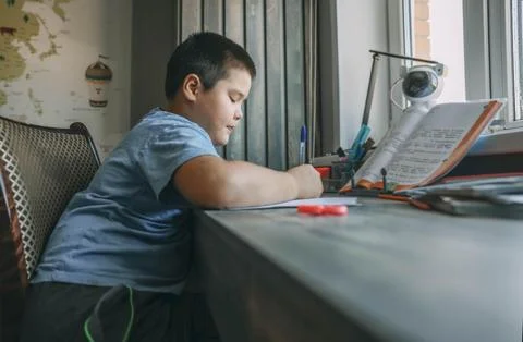 Asian boy write in notebook, homework beside open textbook on desk, study tool Stock Photos
