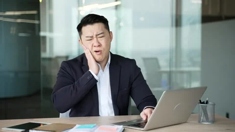 Asian businessman suffering from toothache while sitting at workplace at desk  Stock Footage 321890903