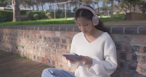 Asian child sitting on brick wall in park with pink headphones tapping tablet, Stock Footage 340676222