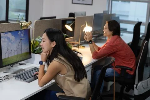 Asian colleagues sitting at open-plan office, woman watching wind turbines on Stock Photos