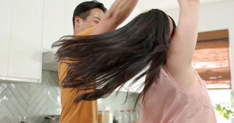Asian couple dancing, man reaching for her hand leading twirl in kitchen by Stock Footage 341932243