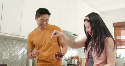Asian couple in kitchen, man offering bowl and spoon, woman drizzling syrup, Stock Footage 335199166