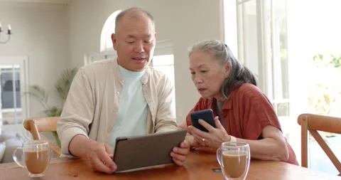 Asian couple sitting at kitchen table, woman showing phone, man moving tablet Stock Footage 342392777
