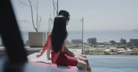 Asian couple sitting at rooftop pool on towel, ocean drawing red dress woman Stock Footage 343516921