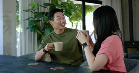 Asian couple sitting at table man raising mug woman lifting hers sipping while Stock Footage 339320924