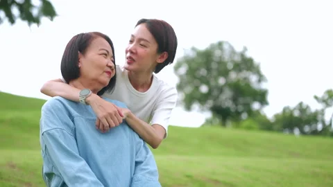 Asian daughter lovingly embraces her mother from behind in a green park. Stock Footage 346509706