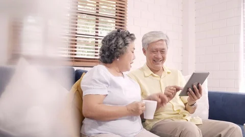Asian elderly couple using tablet and drinking coffee in living room at home, Stock Footage 102522413