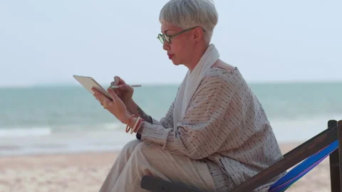 Asian elderly woman using a tablet computer while sitting on the beach. Stock Footage 188020331