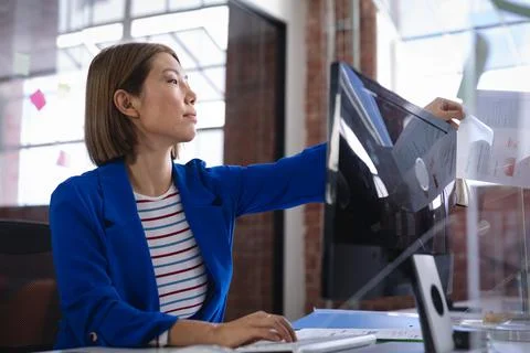 Asian female in front of computers separated by sneeze shield giving document to Stock Photos