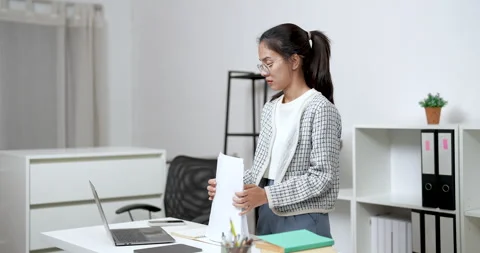Asian female reading printed documents while standing beside desk in modern 库存影片 336377426