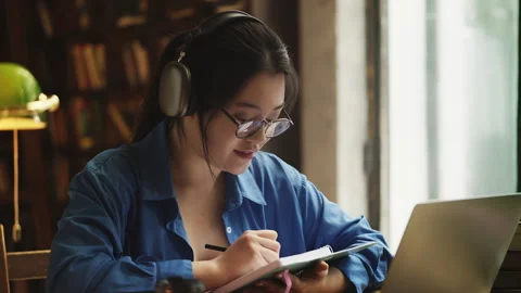 Asian female student in headset having video lesson on laptop, taking notes Stock Footage 331876567