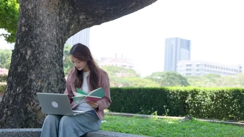 Asian Female Student Using Laptop and Reading Book in City Park Stock Footage 335192457