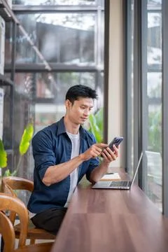 Asian freelancer man checking smartphone beside laptop while sits at counte.. Photos