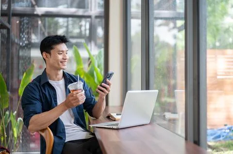 Asian freelancer man drinking iced coffee while using smartphone and laptop Stock Photos