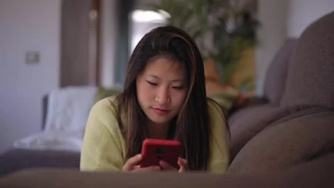 Asian girl lying down on the sofa at home using smartphone spending her free Stock Footage 259592271
