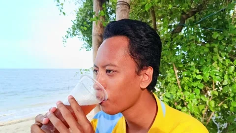 A asian guy is enjoying drinking iced tea with a beach Stock Footage 295477172