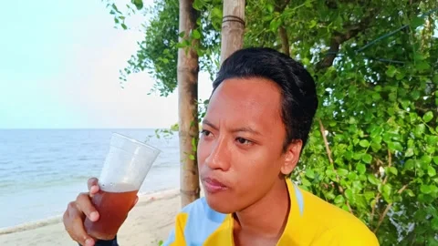 A asian guy is enjoying drinking iced tea with a beach Stock Footage 295477273