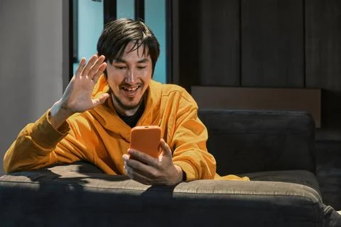 An Asian guy sits on sofa in a small office and communicates by mobile phone Stock Photos