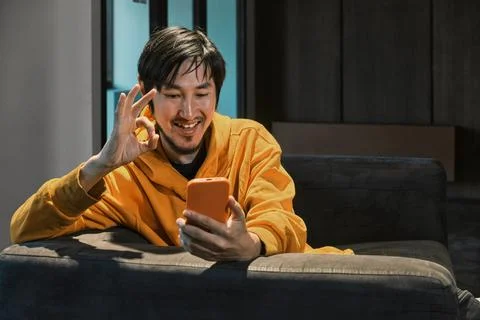 An Asian guy sits on sofa in a small office and communicates by mobile phone Stock Photos