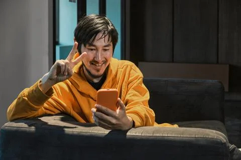 An Asian guy sits on sofa in a small office and communicates by mobile phone Stock Photos
