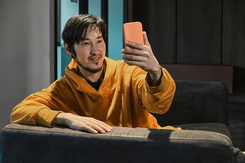 An Asian guy sits on sofa in a small office and communicates by mobile phone Stock Photos