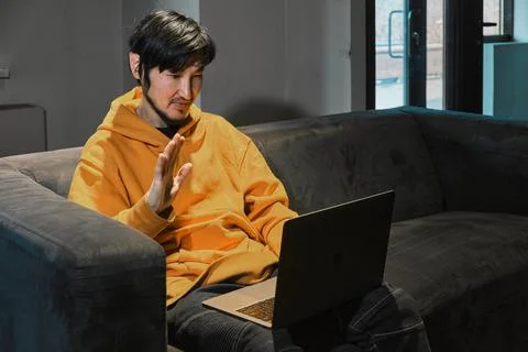 An Asian guy sits on sofa in a small office and communicates via laptop Foto stock