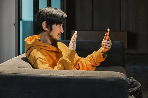 An Asian guy sits on sofa in a small office and communicates by mobile phone Stock Photos