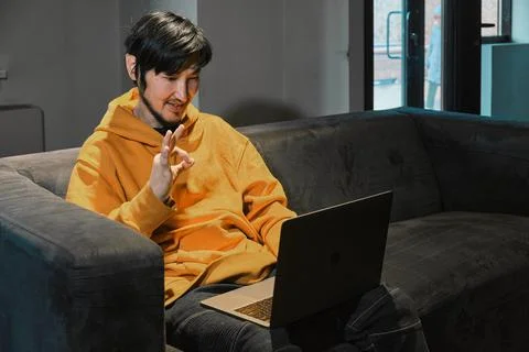 An Asian guy sits on sofa in a small office and communicates via laptop Stock Photos