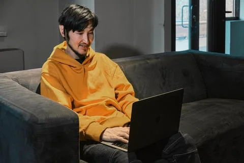 An Asian guy sits on sofa in a small office and communicates via laptop Stock Photos
