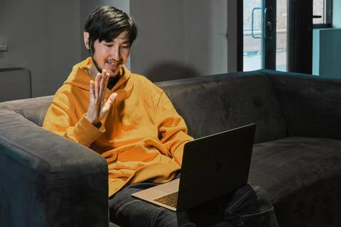 An Asian guy sits on sofa in a small office and communicates via laptop Stock Photos