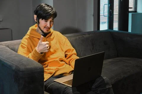 An Asian guy sits on sofa in a small office and communicates via laptop Stockfoto's