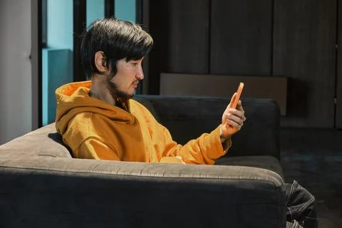 An Asian guy sits on sofa in a small office and communicates by mobile phone Stock Photos