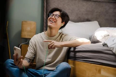 Asian guy using cellphone and earphones while sitting on floor at home Stock Photos