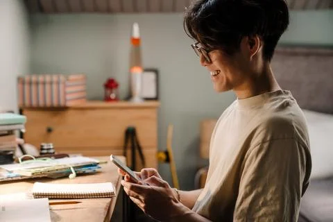 Asian guy using cellphone while sitting at desk and studying at home Stock Photos