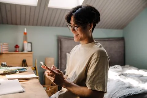 Asian guy using cellphone while sitting at desk and studying at home Stock Photos